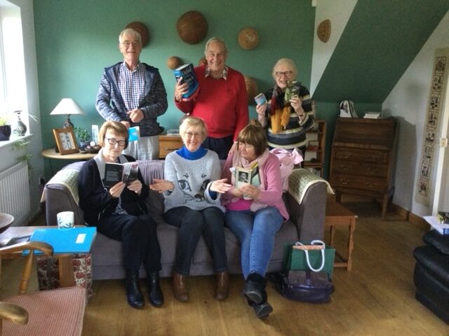 three people sat on a sofa and three stood behind. Holding books and Smiling.